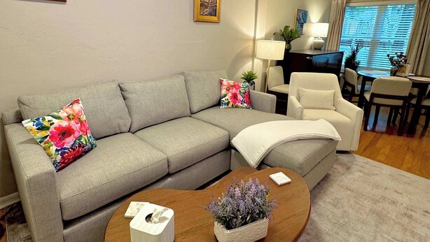 Ein Wohnzimmer mit grauem Sofa, floralen Kissen, einem Couchtisch mit Taschentuchbox und Blumenvase, einem Essbereich mit Stühlen und Tisch sowie einem Deckenventilator.