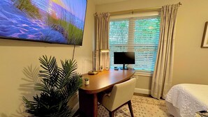 Ein Hotelzimmer mit Schreibtisch, Computer, Fernseher, Bett und einem Fenster mit Vorhängen.