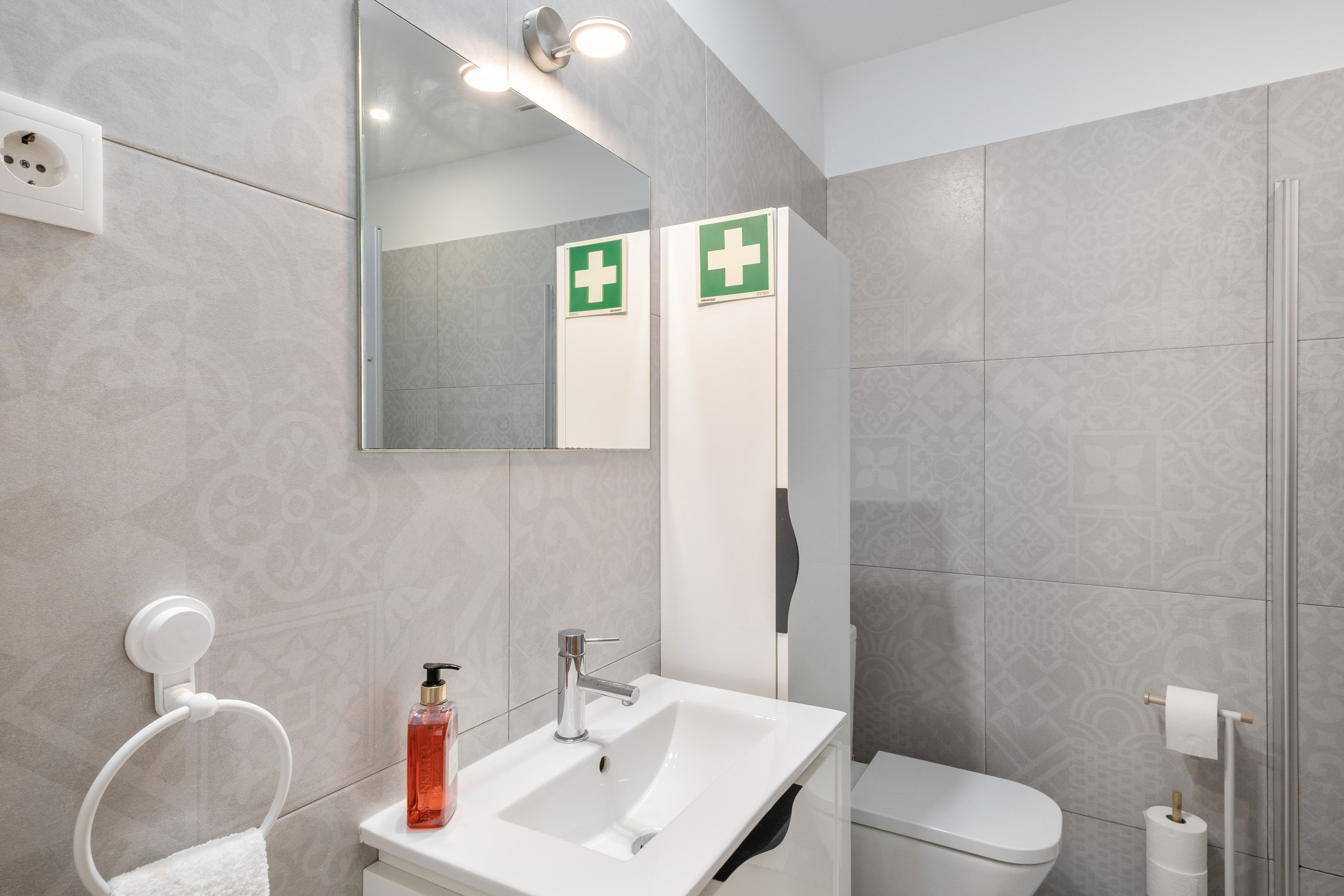 Et moderne badeværelse med en hvid vask, spejl og toilet. Der er to grønne nødskilte på væggen.