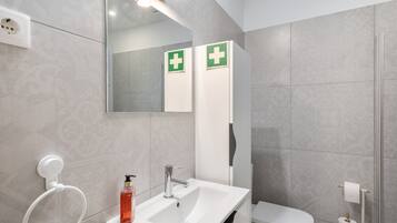 Et moderne badeværelse med en hvid vask, spejl og toilet. Der er to grønne nødskilte på væggen.