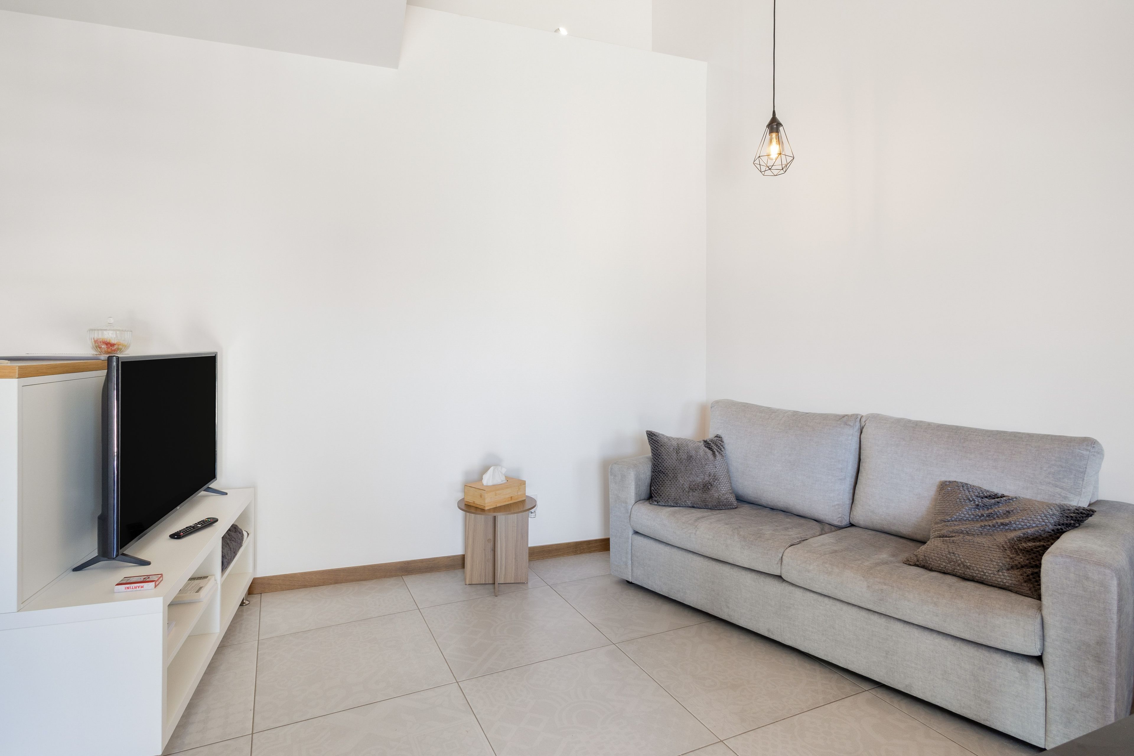 Una sala de estar moderna con un televisor de pantalla plana, un sofá gris con cojines, una mesita de madera y una lámpara colgante.