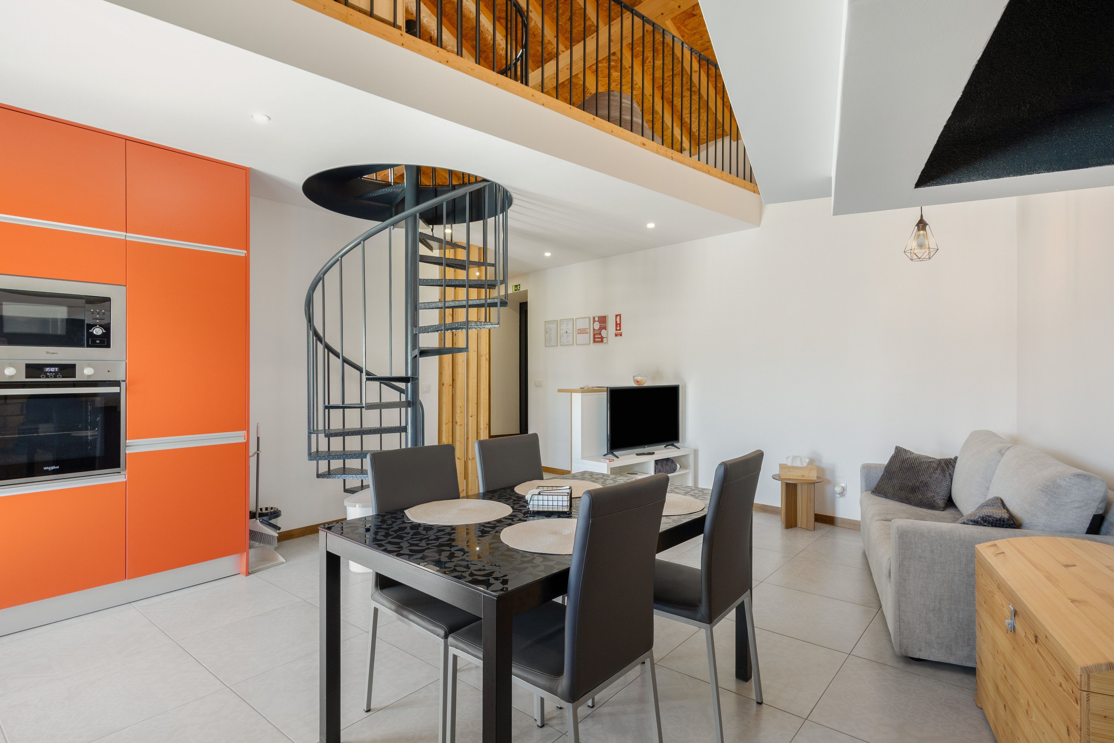 A modern kitchen with a spiral staircase, a dining area with a TV, and a living room with a sofa and a wooden coffee table.