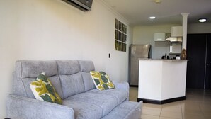 Ein Wohnzimmer mit grauem Sofa, zwei gemusterten Kissen, einem Glastisch und einer Küchenzeile im Hintergrund mit Kühlschrank und Herd.