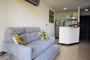 Ein Wohnzimmer mit grauem Sofa, zwei gemusterten Kissen, einem Glastisch und einer Küchenzeile im Hintergrund mit Kühlschrank und Herd.
