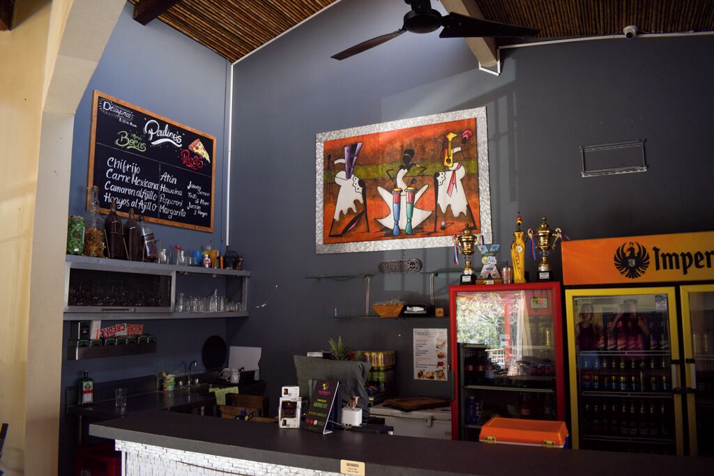 Un coin bar avec un tableau noir affichant le menu, où l’on peut trouver des plats comme des « Chiles Rellenos » et de la « Carne Mexicana ». Un mur est décoré d’une fresque représentant des musiciens, et un réfrigérateur est rempli de boissons.