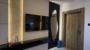 A modern bedroom with a large bed, a TV mounted on the wall, a wooden door, and a small trash bin. - BAE LUXURY APARTMENT (Akure)