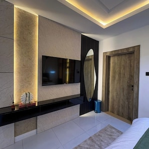 A modern bedroom with a large bed, a TV mounted on the wall, a wooden door, and a small trash bin. - BAE LUXURY APARTMENT (Akure)