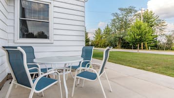 En terrasse med et rundt bord og fire stole, et hvidt hus med et vindue, og et grønt område med træer.