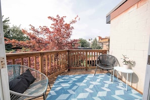 Ein Balkon mit Holzgeländer, zwei Korbstühle, einem kleinen Tisch und einem blau gemusterten Teppich.