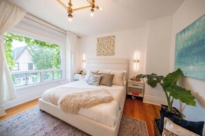 A bedroom with a large bed, a window with curtains, a chandelier, a nightstand, a wall art piece, and a potted plant. - Charming Victorian Home | Little Italy + Garden (Toronto)
