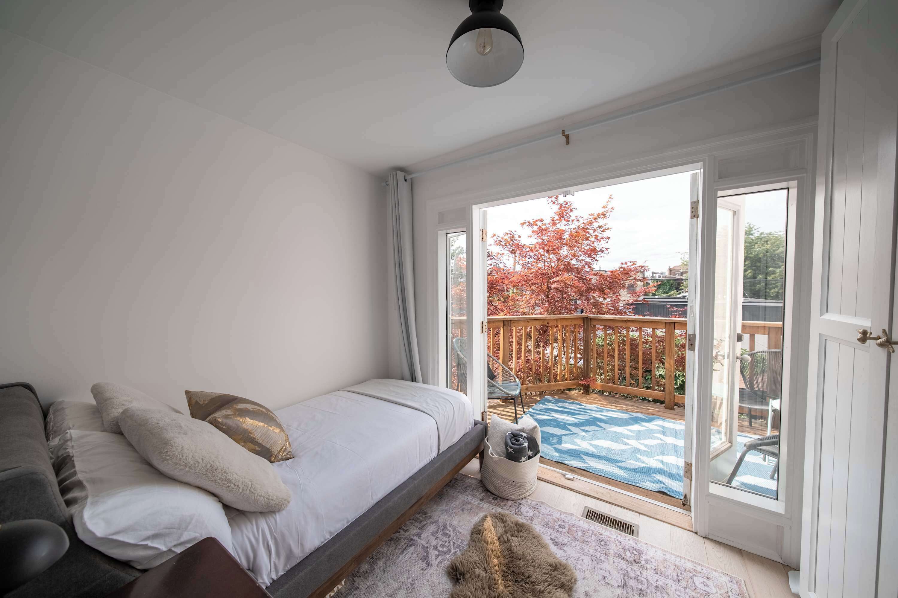 Una camera da letto con un letto, una sedia e vista su un balcone con pavimento in legno e foglie autunnali.