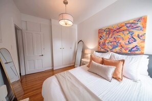 Ein Schlafzimmer mit einem Bett, einem Nachttisch, einer Lampe, einem Spiegel und einem farbenfrohen, abstrakten Gemälde an der Wand.