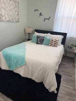 A bedroom with a bed, pillows, a wall art piece, a bedside lamp, and a window with blinds. - SMOKE FRIENDLY Lovely 1-bedroom apartment with AC in vibrant Racine (Racine)