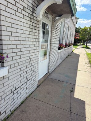 A white brick building with a numbered door, a window with a flower box, and a sidewalk. - SMOKE FRIENDLY Lovely 1-bedroom apartment with AC in vibrant Racine (Racine)