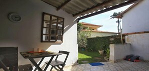 A room with a table and chairs, a clock on the wall, and a view of a garden and neighboring buildings. - Casinha de Hóspede em Búzios ao lado Orla Bardot (Buzios)