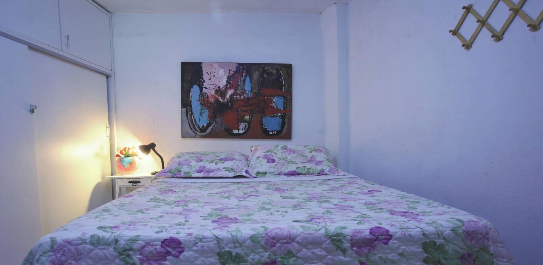 Un dormitorio con cama, mesita de noche, lámpara y un cuadro en la pared.