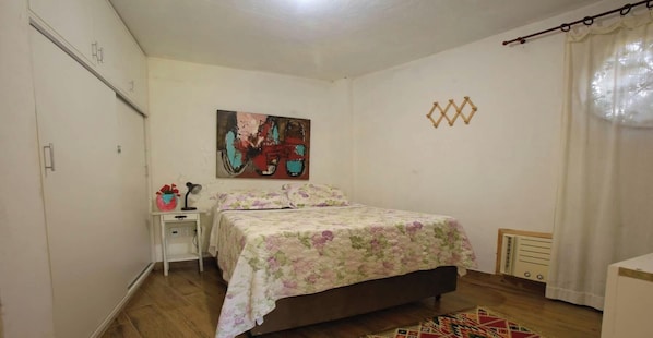 A bedroom with a bed, a nightstand, a painting, and a radiator. - Casinha de Hóspede em Búzios ao lado Orla Bardot (Buzios)