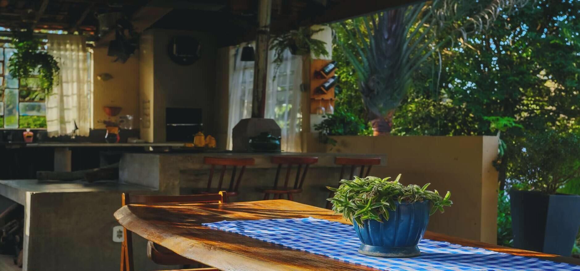 A wooden table with a blue and white checkered tablecloth, a potted plant, and a view of a kitchen area with chairs and a counter.