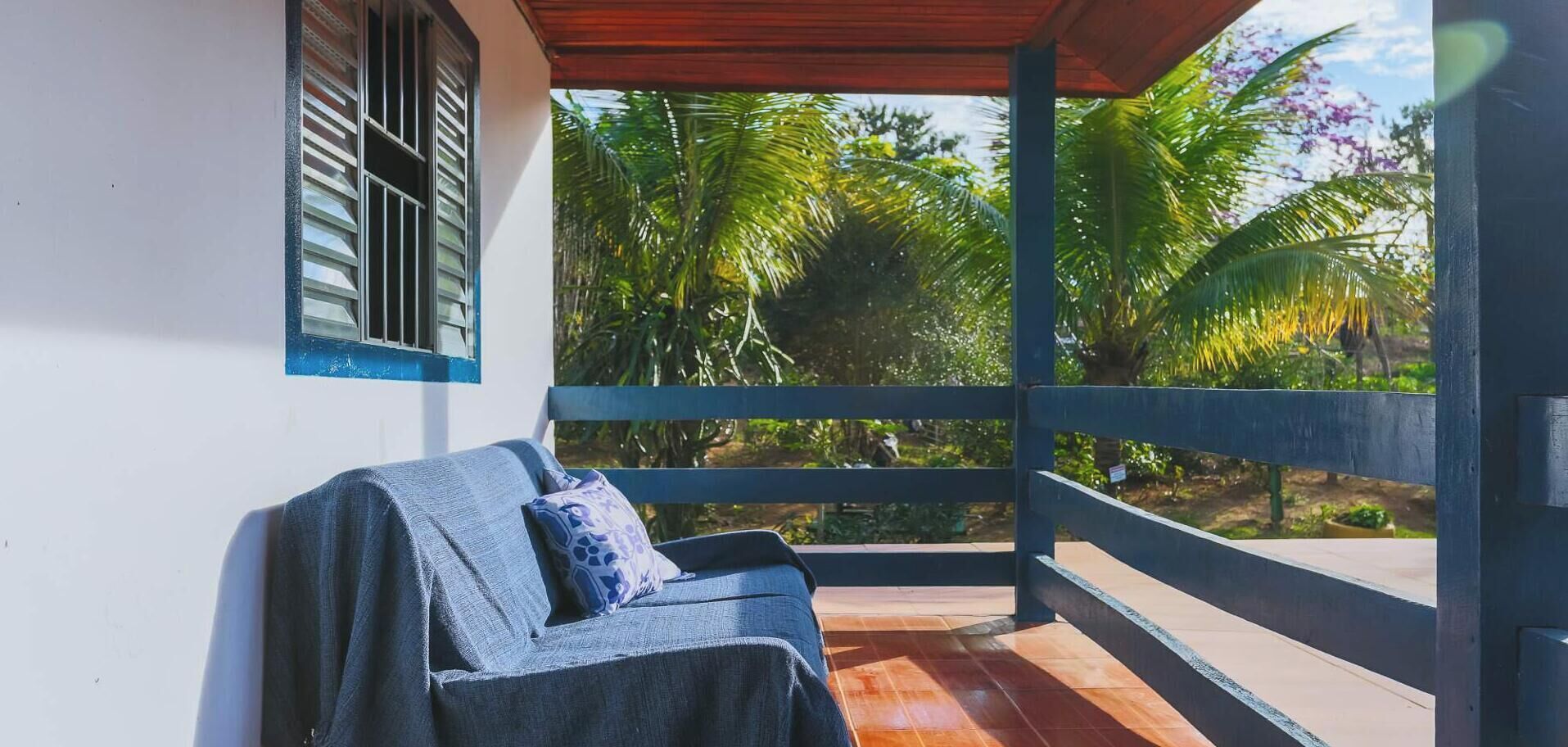 A covered outdoor seating area with a bench, a window with shutters, and a view of tropical vegetation.