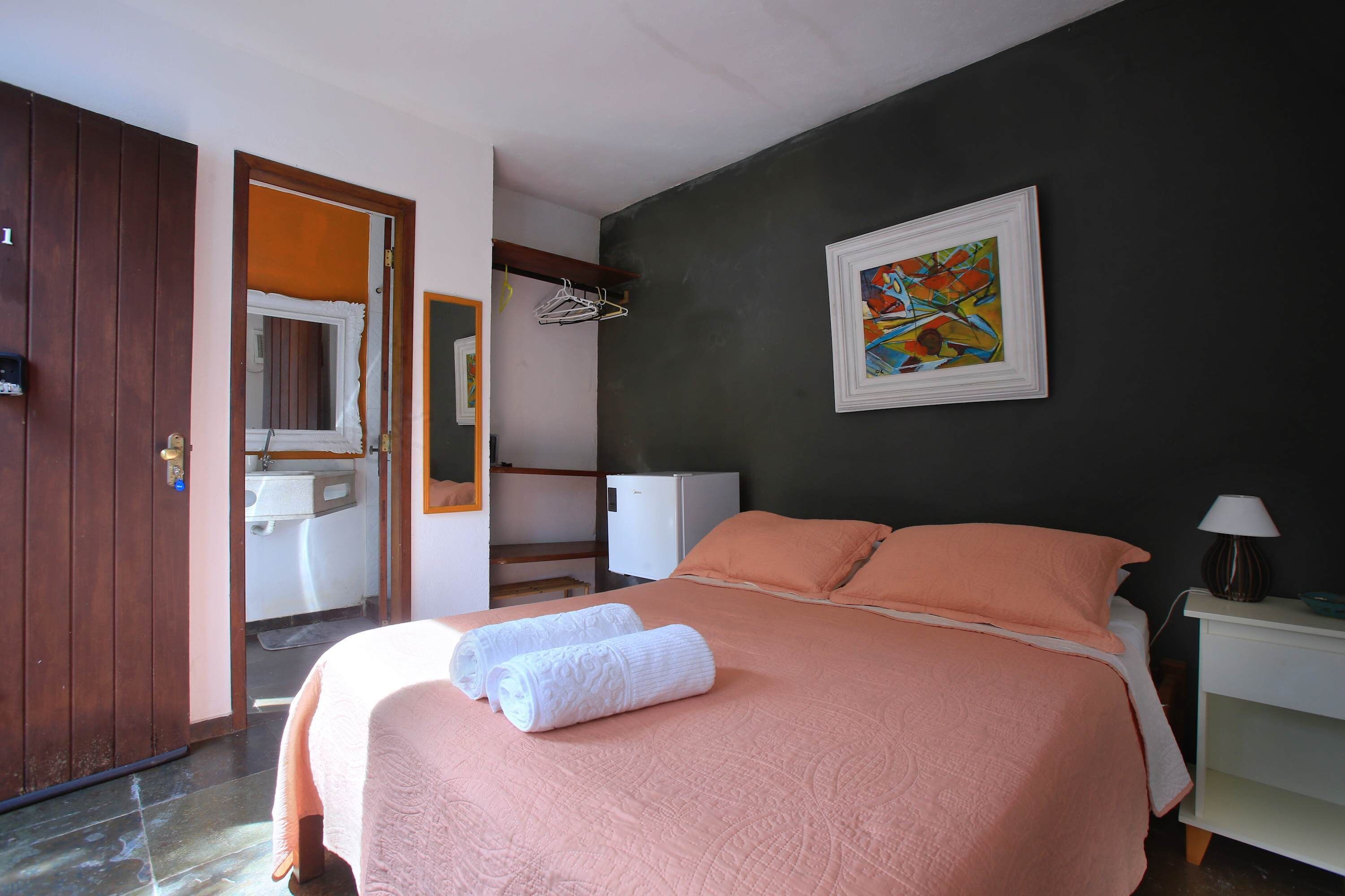 A bedroom with a bed, a nightstand, a lamp, a painting, a bathroom with a mirror, and a door.