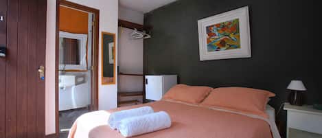 Un dormitorio con cama, mesita de noche, lámpara, un cuadro, un baño con espejo y una puerta.