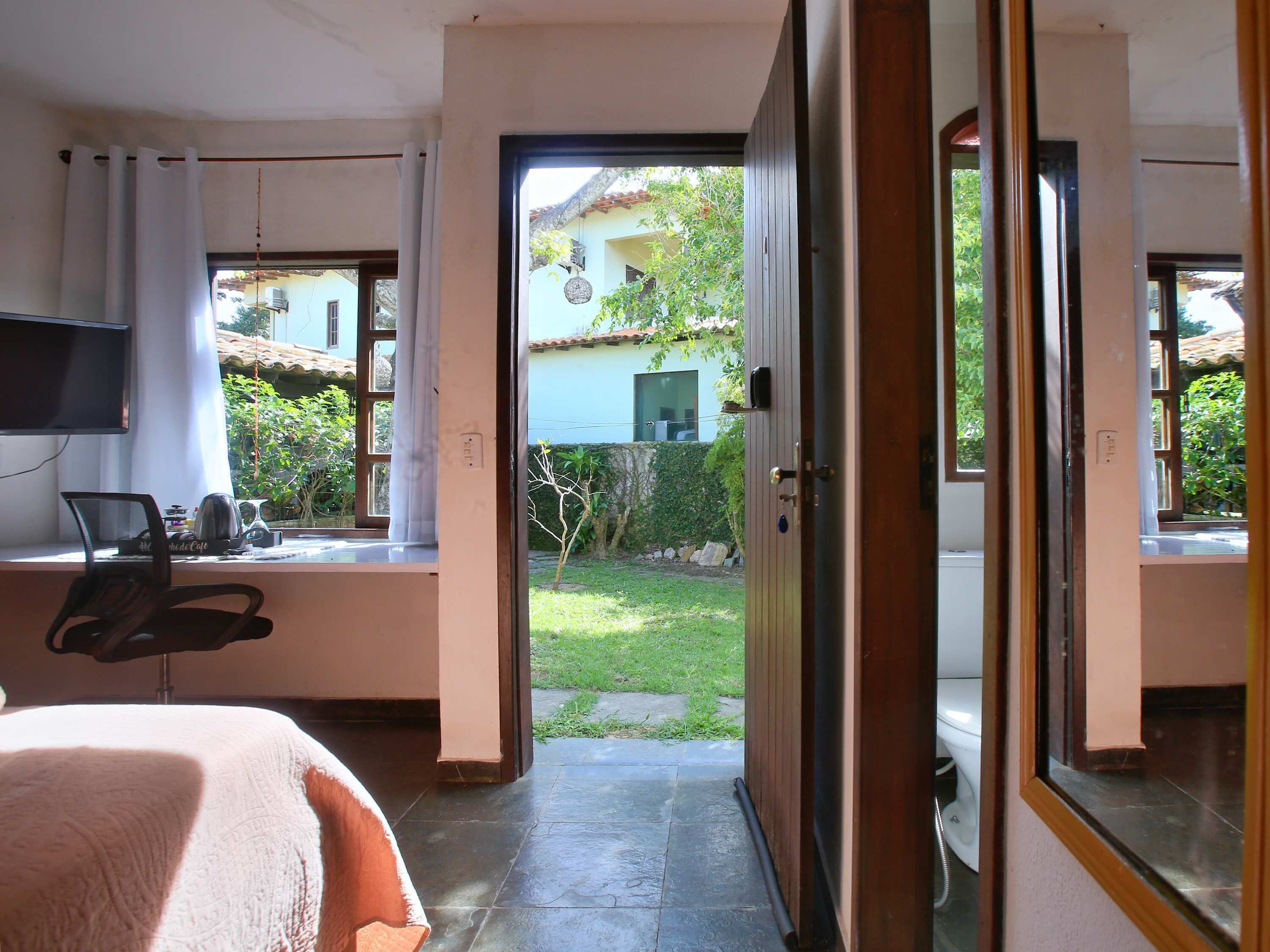 A room with a bed, a desk with a chair, a television mounted on the wall, and a door leading to an outdoor area with a view of a house and greenery.