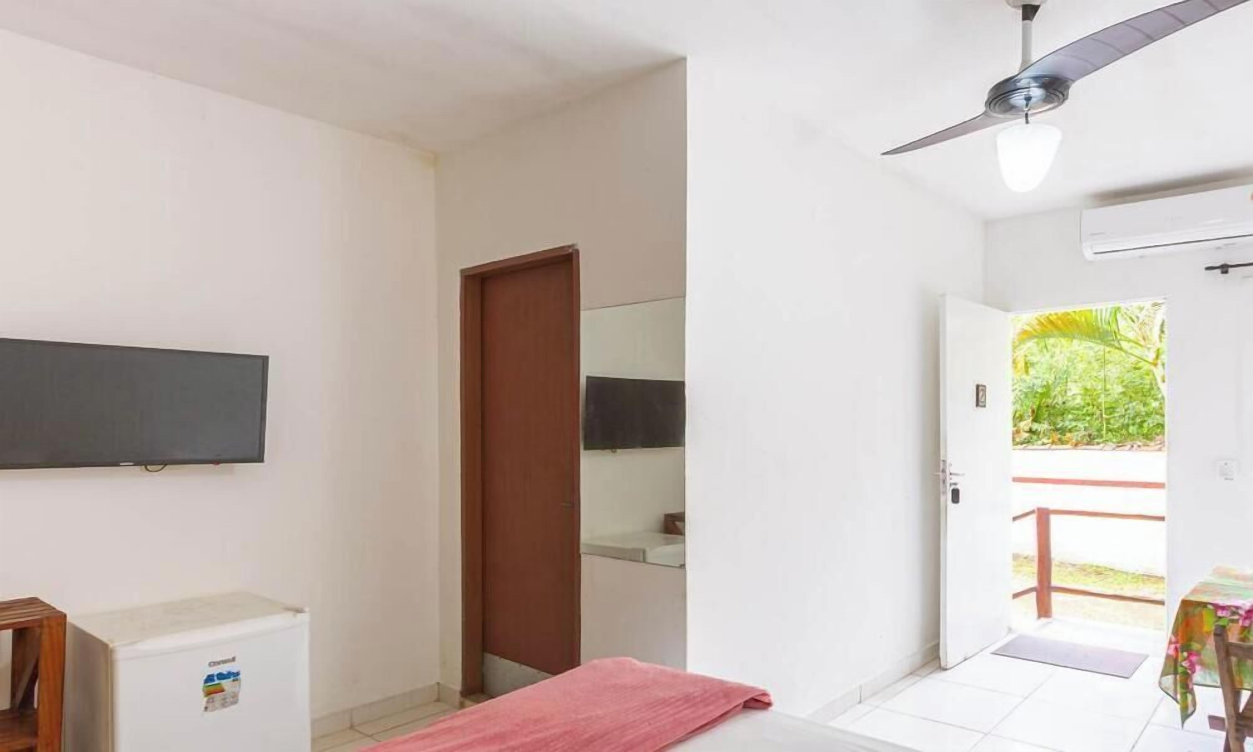 A room with a ceiling fan, a flat-screen TV mounted on the wall, a small refrigerator, and a door leading to an outdoor area with a view of greenery.