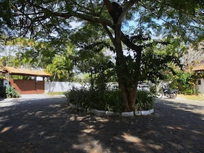 A paved courtyard with a tree in the center, surrounded by a low white wall and a brick building with a tiled roof. - Suíte Dupla em Buzios Perto da Rua das Pedras (Armação dos Búzios)