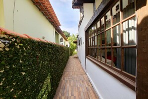 A narrow pathway flanked by a hedge and a building with large windows. - Suíte Dupla em Buzios Perto da Rua das Pedras (Armação dos Búzios)