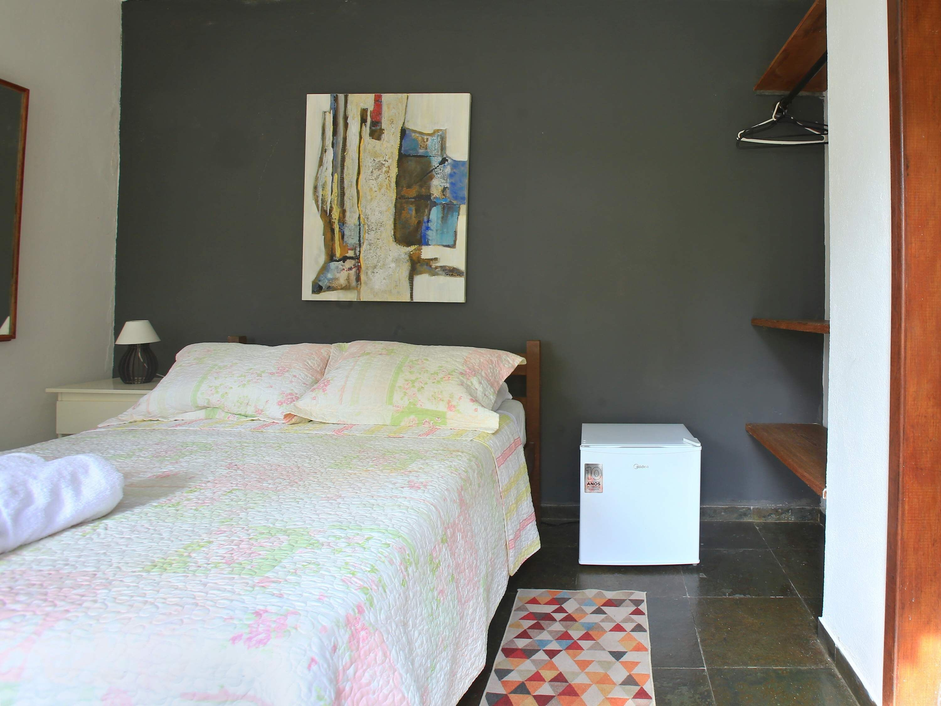 A bedroom with a bed, a nightstand, a lamp, a refrigerator, a geometric rug, and a painting on the wall.