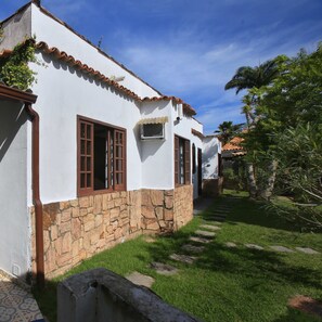 A white building with stone foundation, a tiled roof, and a paved pathway leading to a door. - Suíte Dupla em Buzios Perto da Rua das Pedras (Armação dos Búzios)