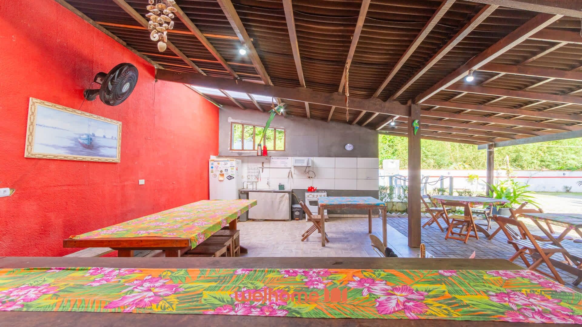 A covered outdoor dining area with a red wall, a kitchen, and a view of greenery.