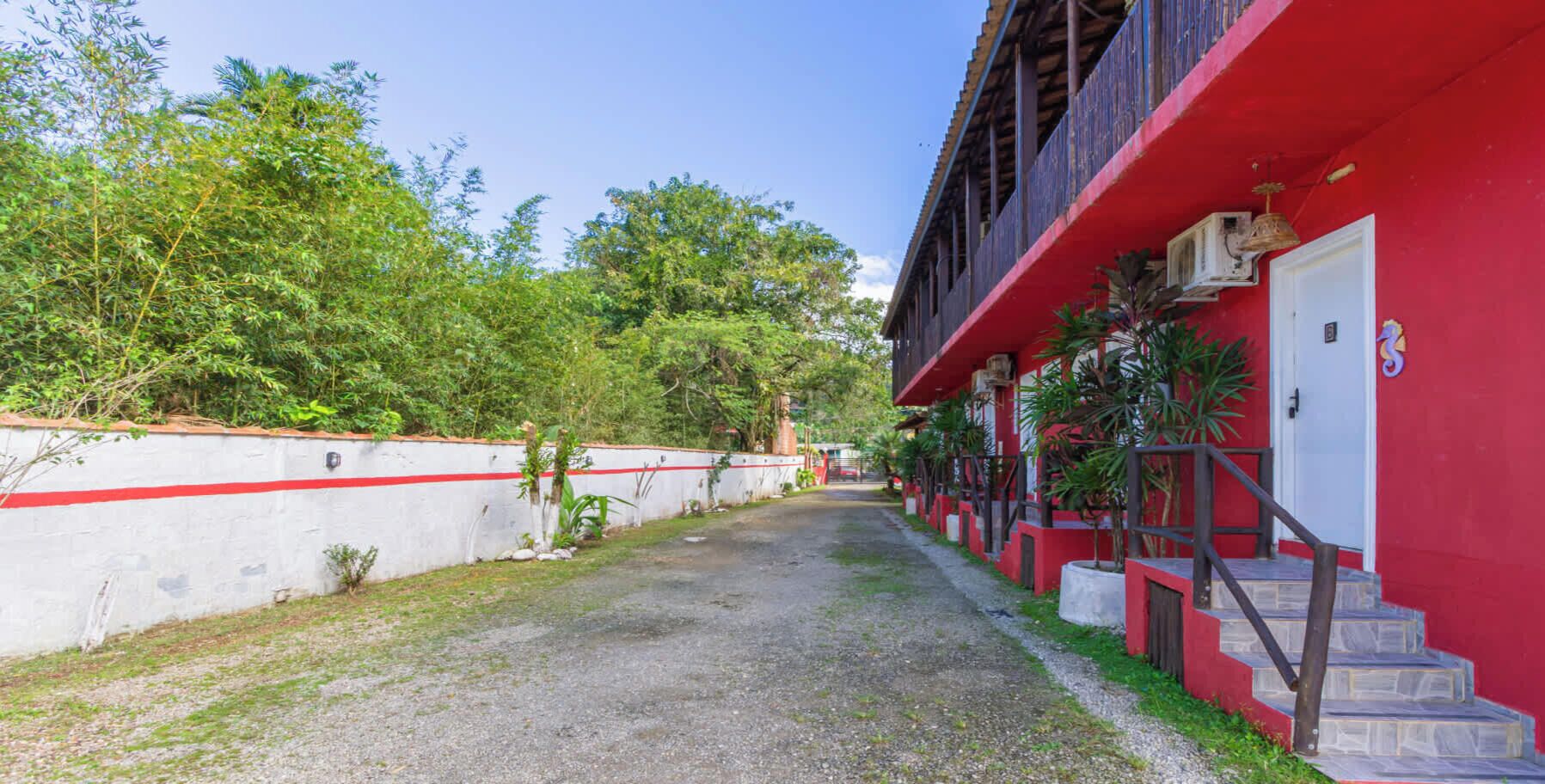 Ein Weg führt zu einem Gebäude mit roter Fassade, einer weißen Wand mit rotem Streifen und einer Treppe mit roter Geländer.