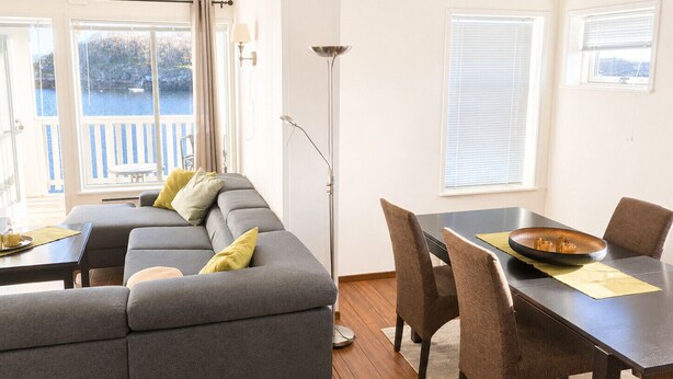 A modern living room with a grey sectional sofa, a dining table with chairs, and a view of the outdoors through large windows. - Apartment with sea view in Henningsvær (Vågan)