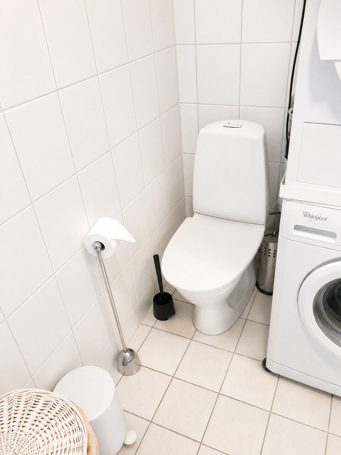 A small, clean bathroom with a toilet, a bidet, a washing machine, and a trash bin.