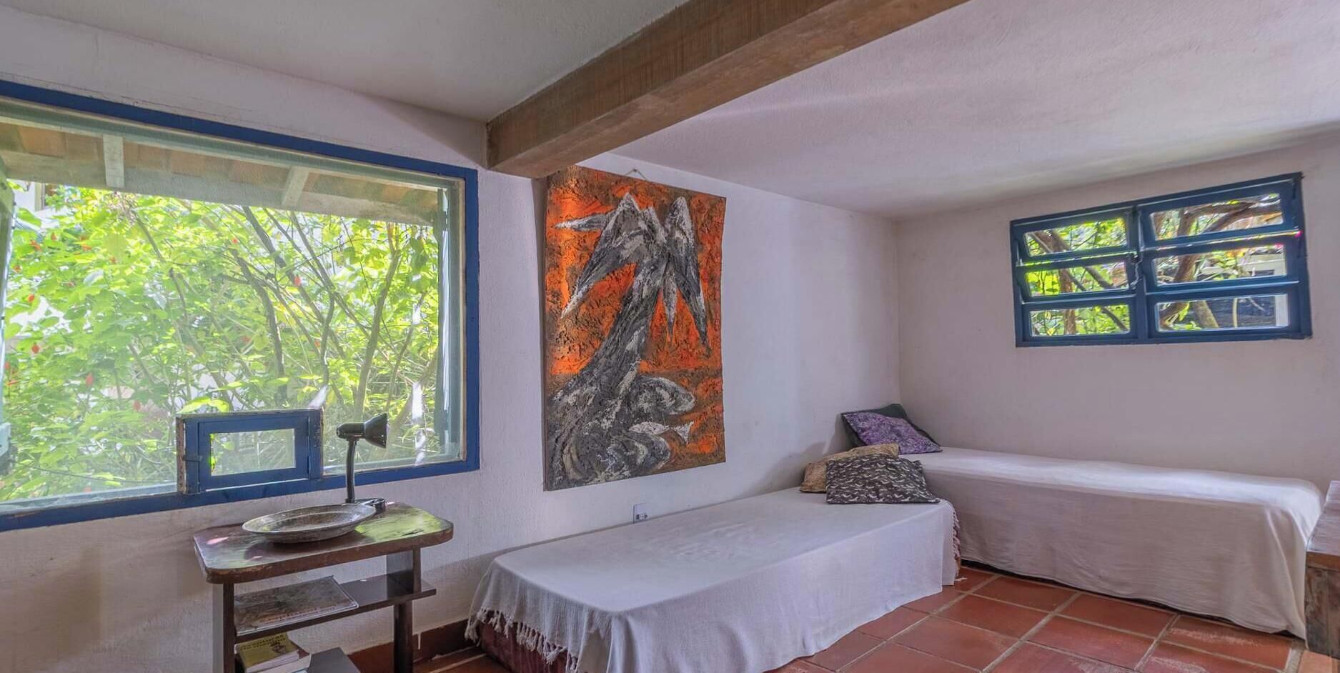 A room with a bed, a table with a lamp, a painting, and a window with a view of trees.
