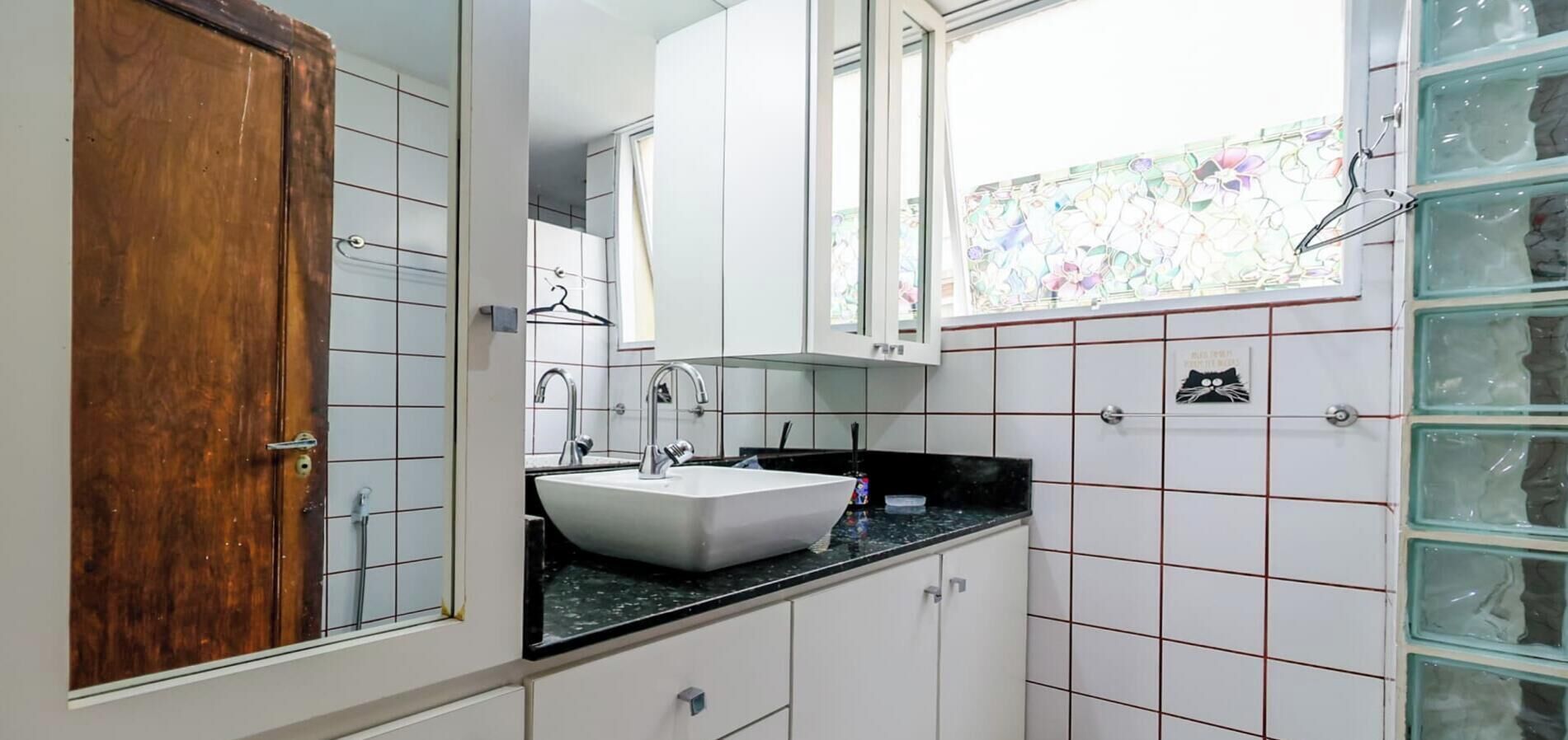 A bathroom with a wooden door, a white sink, a mirror, and a window with a floral view.