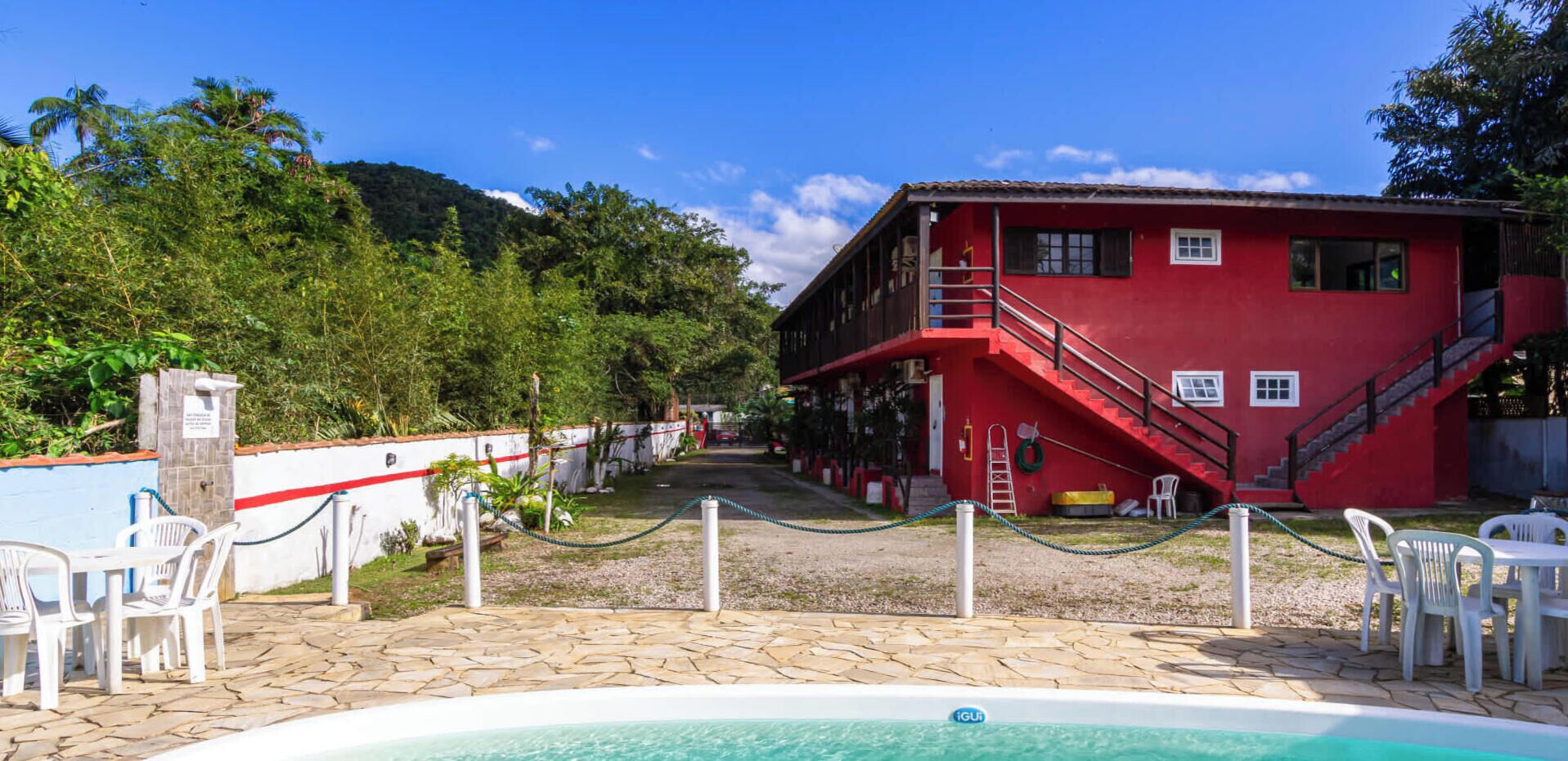 Un bâtiment rouge doté d’un espace piscine, entouré de verdure et surplombé d’un ciel dégagé.