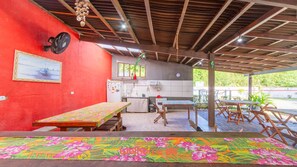 Un espace extérieur abrité, aménagé pour les repas, avec un mur rouge, une cuisine et une vue sur la verdure.