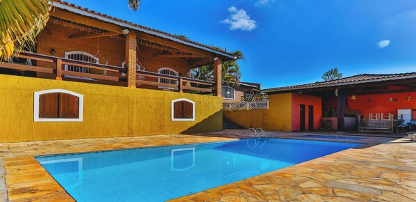 A pool area with a clear blue pool, a yellow wall with a window, and a red building in the background. - Sitio em Atibaia com piscina e churrasqueira (Atibaia)