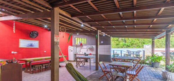 A covered outdoor area with tables and chairs, a red wall, and a kitchen area. - Suite com Piscina a 400m da Praia de Itamambuca (Ubatuba)