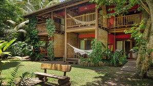 Une maison en bois à deux étages, dotée d’un hamac, d’un banc en bois et d’un jardin où poussent différentes espèces de plantes.