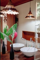 Baño con dos lavamanos blancos, un espejo y una cómoda de madera. Hay dos jarrones negros con hojas verdes y una flor roja sobre la cómoda.