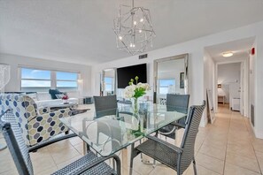 A modern living room with a glass dining table, wicker chairs, a sofa with patterned cushions, a large mirror, and a geometric chandelier. - Million Dollar Gulf View-2 Bed 2 Bath (Clearwater)