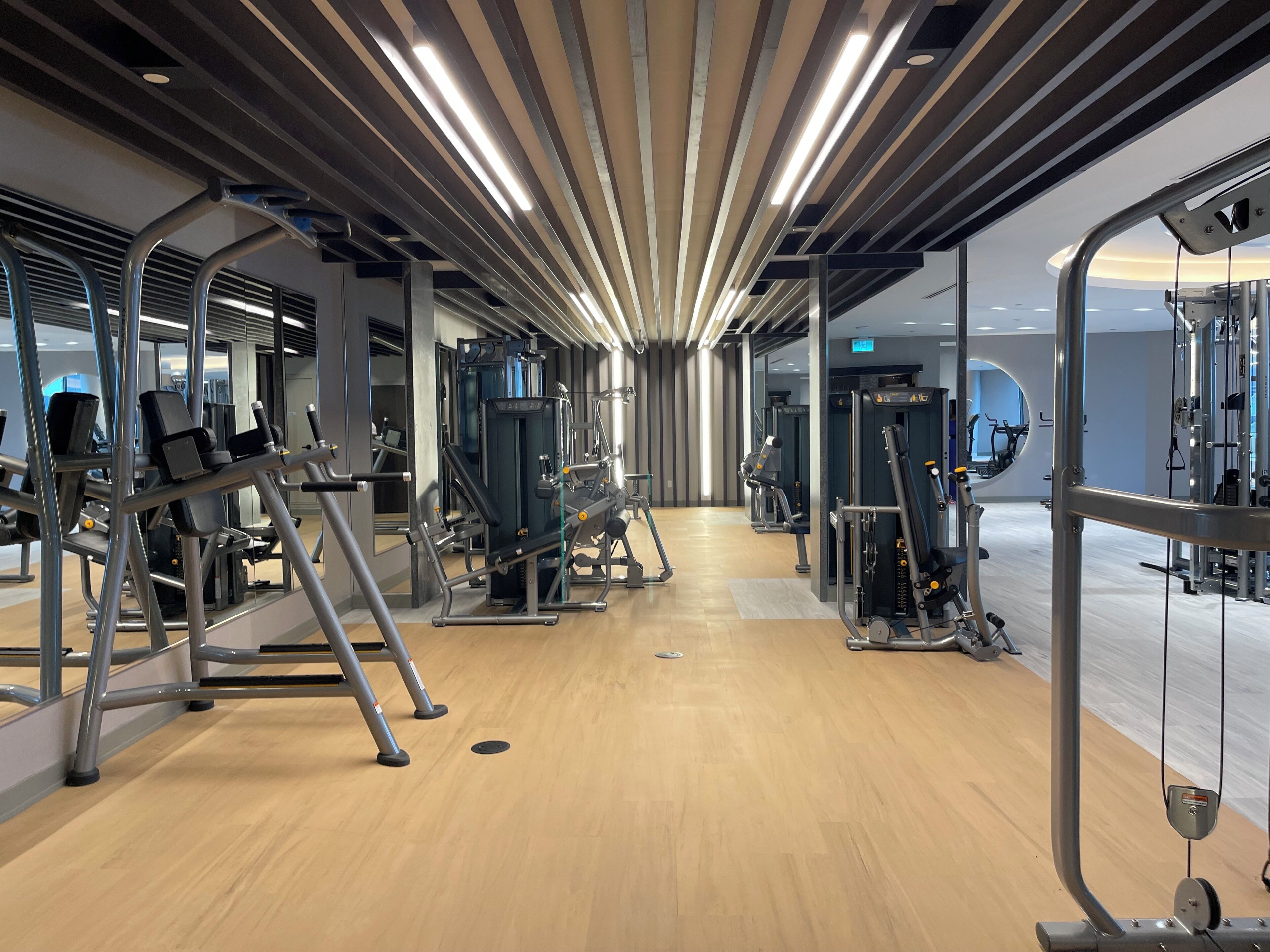 Un gimnasio moderno con diversas máquinas de ejercicio, piso de madera y techo con luces lineales.