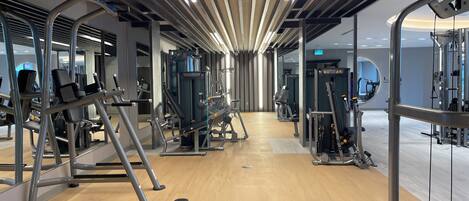 A modern gym with various exercise machines, a wooden floor, and a ceiling with linear lights.