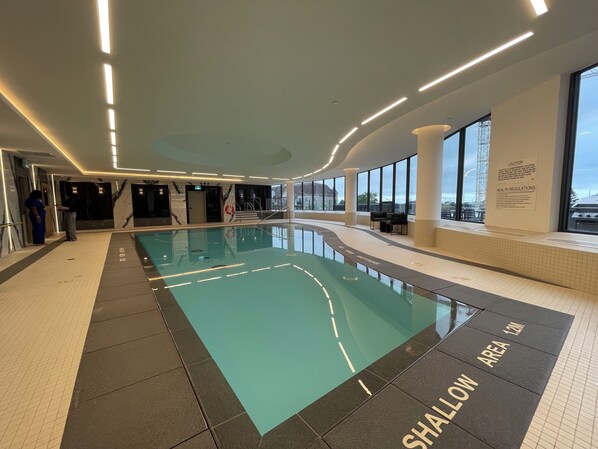 Una piscina interior con zona poco profunda, rodeada de suelo de baldosas y grandes ventanales con vistas al exterior.