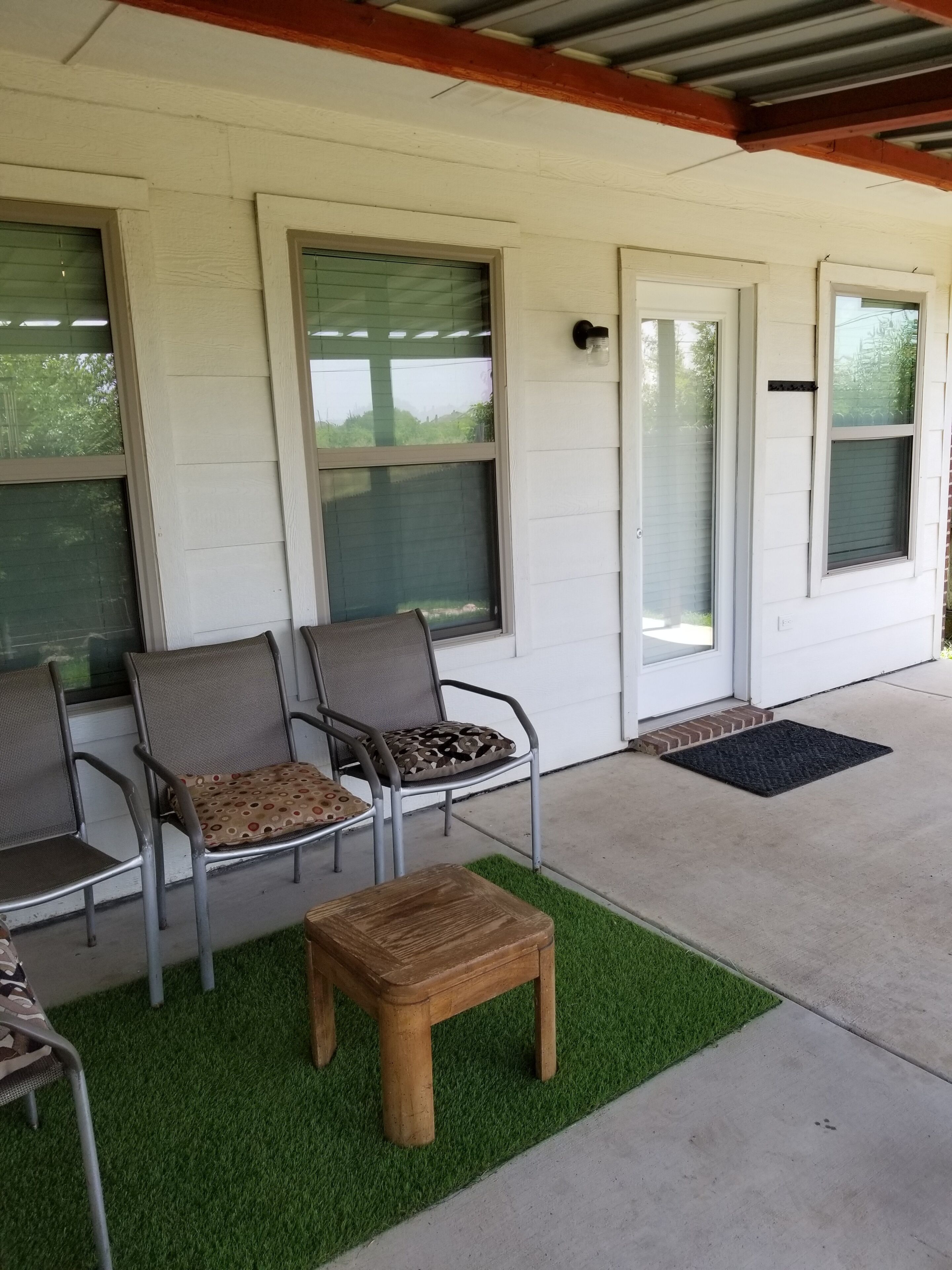 Un espace extérieur abrité, aménagé avec des chaises et une petite table, une porte et une fenêtre donnant sur un paysage verdoyant.