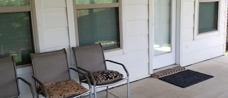 Un espace extérieur abrité, aménagé avec des chaises et une petite table, une porte et une fenêtre donnant sur un paysage verdoyant.