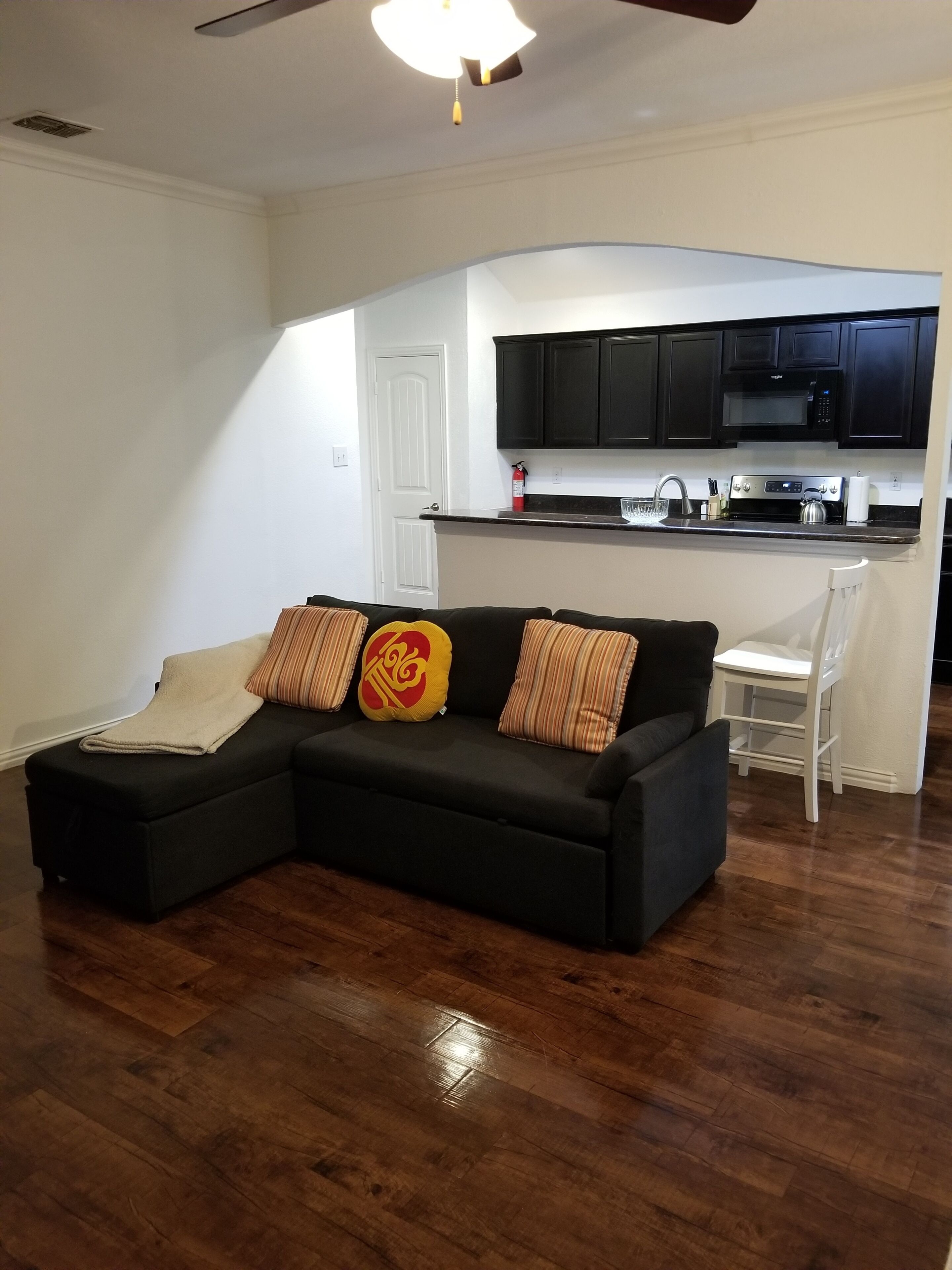 A living room with a sectional sofa, a kitchen with dark cabinets, and a white door.